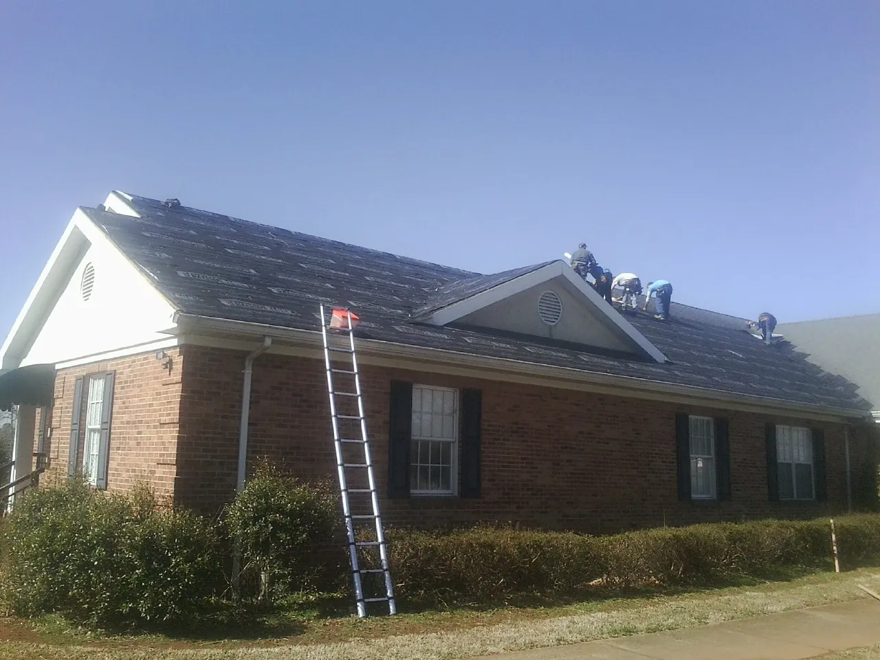 Crew tearing shingles off a brick ranch roof with a ladder set — Emergency Roof Repair work by our crew in Hume