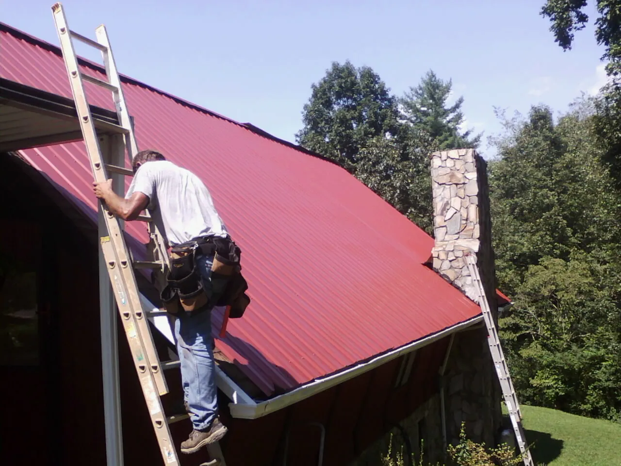Roofer on a ladder beside a red metal roof and stone chimney — Soffit & Fascia Repair by Roofing Brothers in Hume